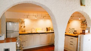 Una cocina con armarios blancos, un lavamanos doble y un refrigerador. Hay luces colgantes y una estantería con utensilios de cocina.