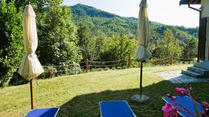 Un jardín con dos sillones azules, dos sombrillas beige y vistas a una montaña boscosa.