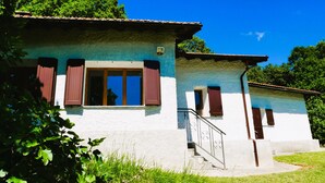 Una casa de dos pisos con fachada blanca y contraventanas marrones, una escalera visible y un cielo azul despejado al fondo.