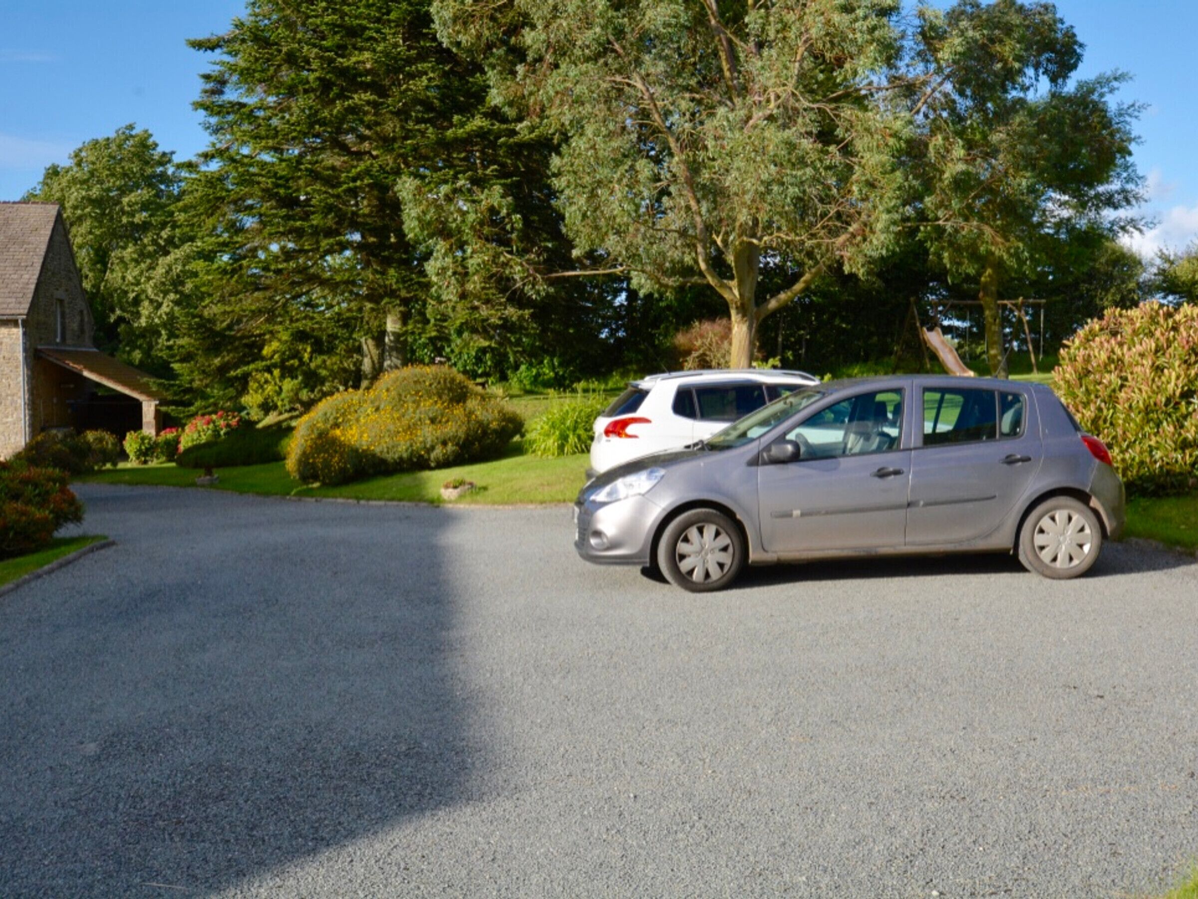 A residential driveway with a parked silver car and a white car. There is a stone building with a sloped roof on the left.