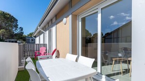 En uteplats med ett vitt bord och stolar, en glasdörr som leder ut till en trädgård och en liten trädgårdsyta med konstgräs.