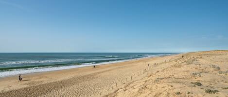 A wide sandy beach with gentle waves, a clear sky, and sparse vegetation.