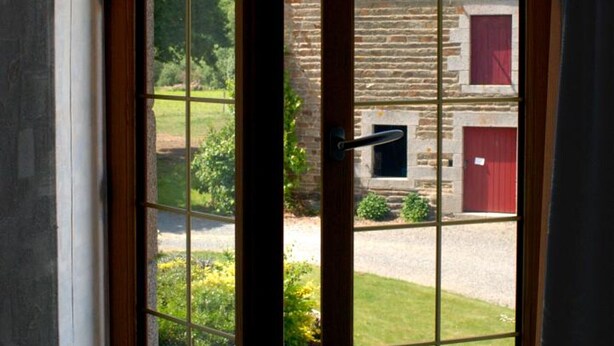 A view through a window with a wooden frame, looking out to a stone building with a red door and a small window. - Charming house in Vessey with TV and internet (Pontorson)