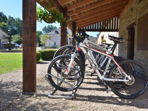 A covered bike parking area with a gravel ground, a wooden pergola, and a stone building in the background.