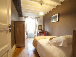 A bedroom with a wooden wardrobe, a bed with white bedding, a desk with a chair, and a painting on the wall.