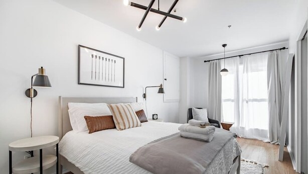 Ein modernes Schlafzimmer mit einem großen Bett, Nachttischen, einem gerahmten Bild an der Wand und einem Fenster mit Vorhängen.