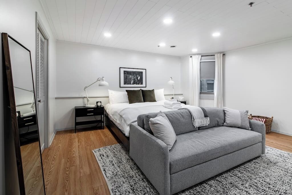 A modern bedroom with a bed, bedside tables, a grey sofa, and a large window.