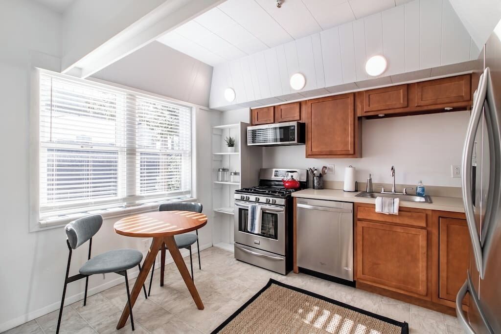 A modern kitchen with wooden cabinets, stainless steel appliances, a dining area with a round table and chairs, and a window with blinds.