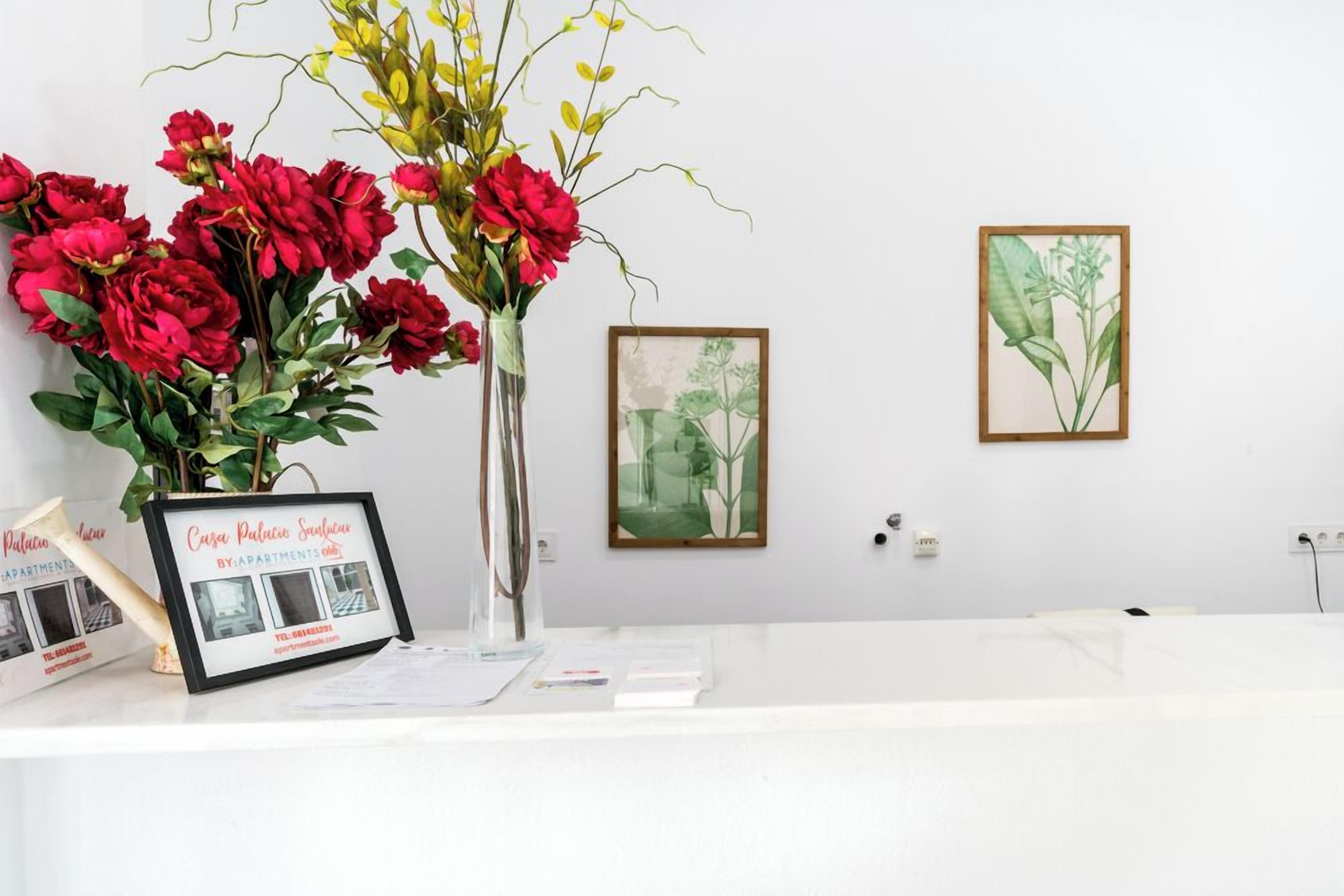 A reception desk with a framed sign, a vase of red flowers, and two framed botanical illustrations on the wall.