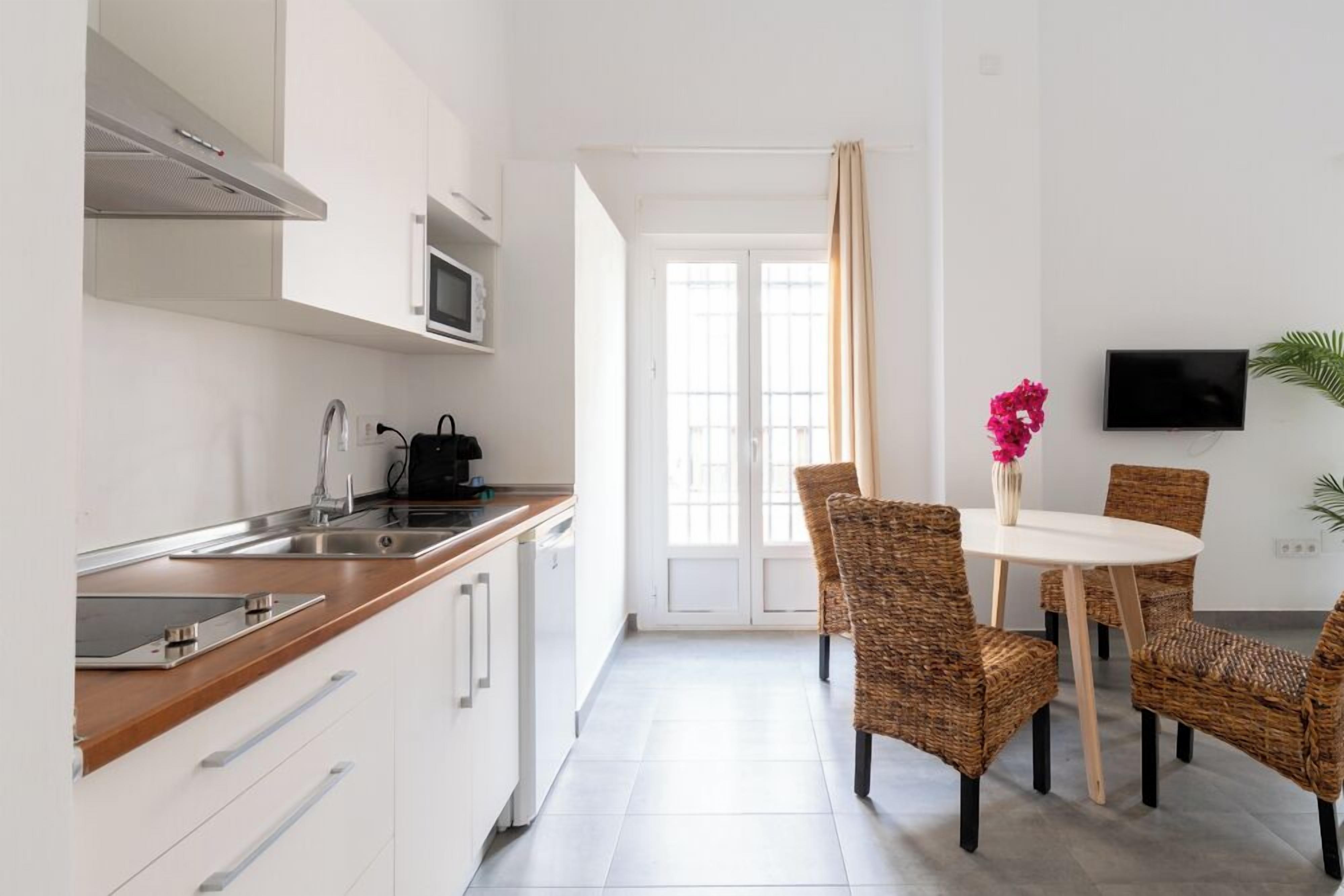 Een moderne keuken met witte kastjes, een roestvrijstalen spoelbak en een magnetron. Er is een eethoek met rieten stoelen en een witte tafel.