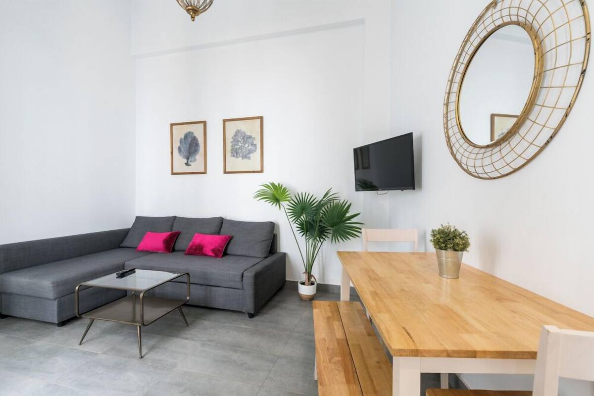 A modern living room with a grey sofa, a wooden dining table, a TV, and a large mirror.