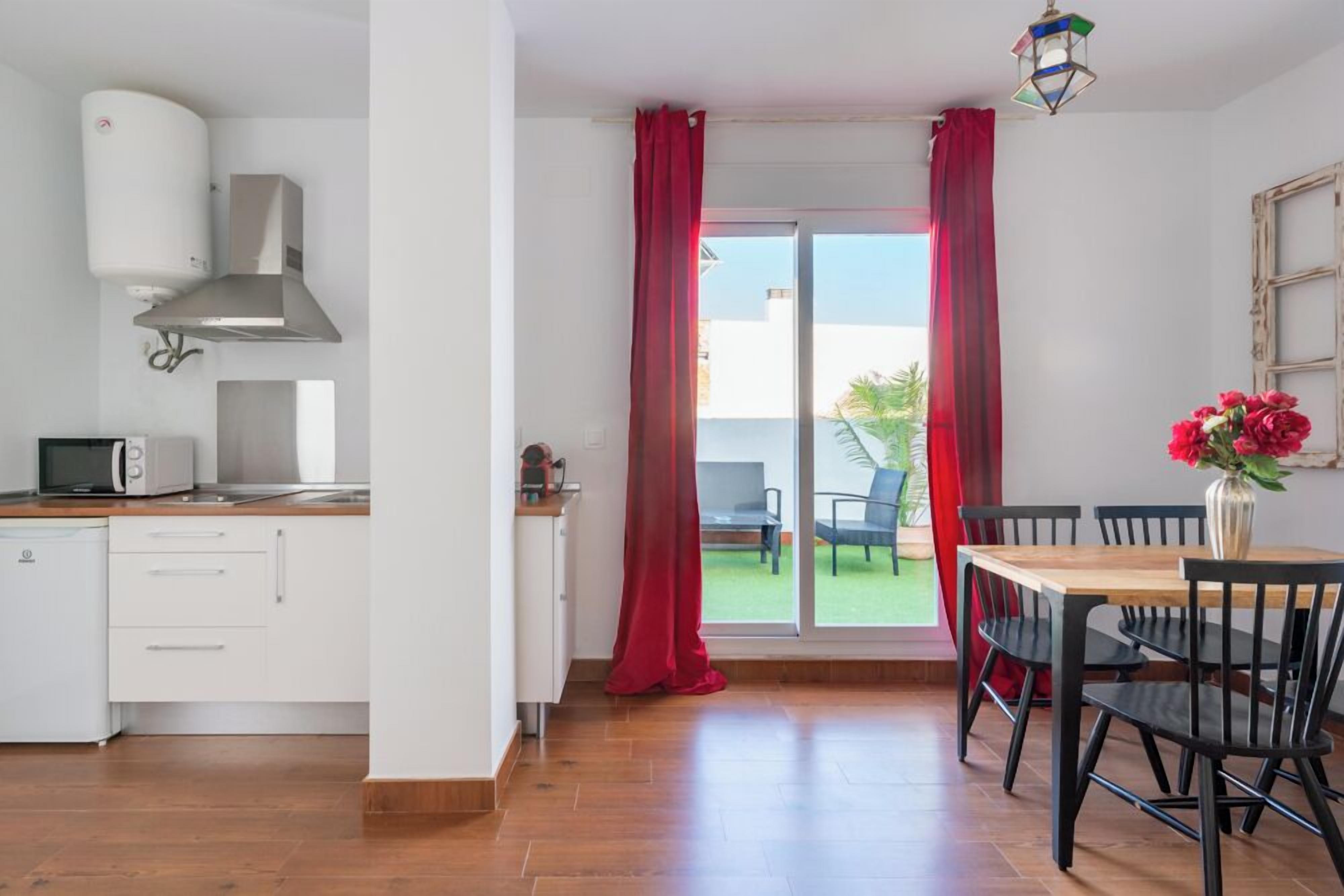 A modern kitchen with white cabinets, a microwave, and a stove. There is a dining area with a wooden table and black chairs. Red curtains cover the sliding glass door leading to a balcony with a chair and a plant.