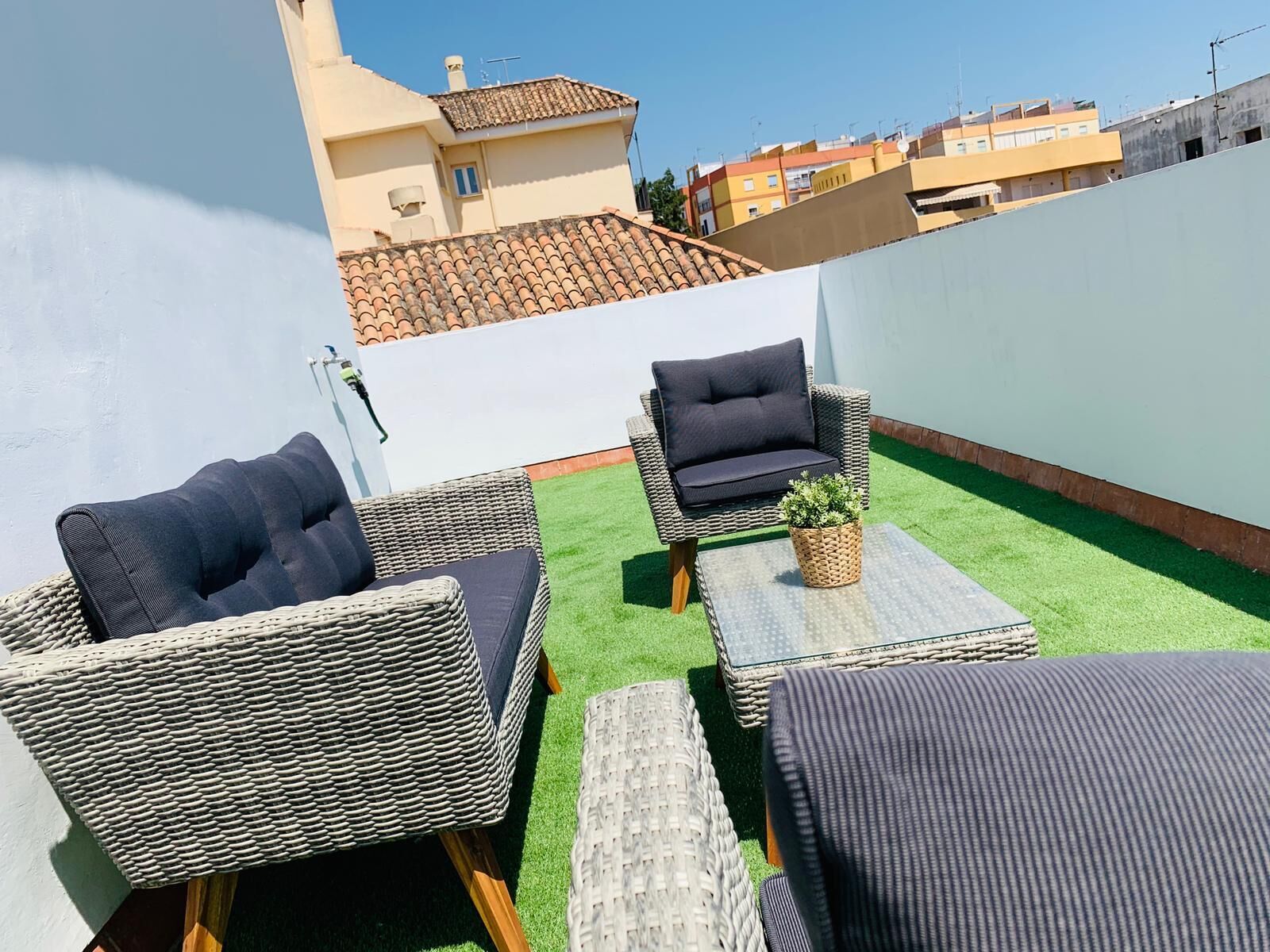 Terraza en la azotea con mobiliario de mimbre, una pequeña mesa con una planta y vistas de edificios circundantes.