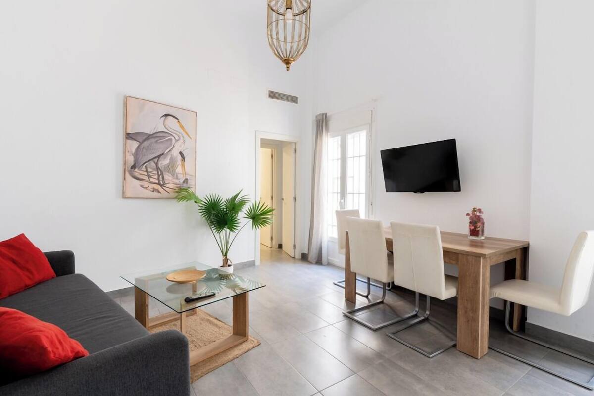 A modern living room with a sofa, a glass coffee table, a dining table with chairs, a potted plant, and a mounted TV.