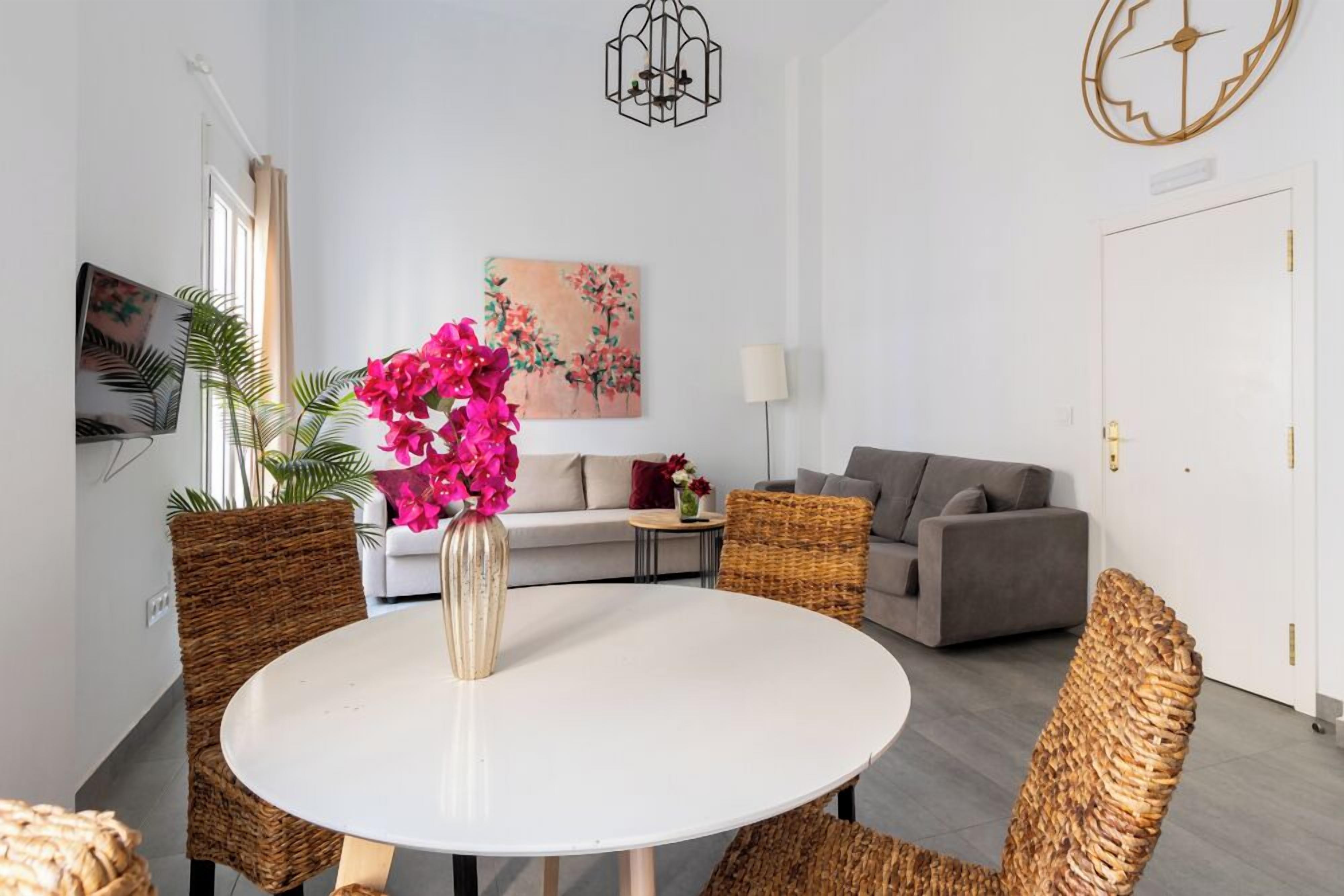 A modern living room with a round dining table, wicker chairs, a grey sofa, and a painting on the wall.