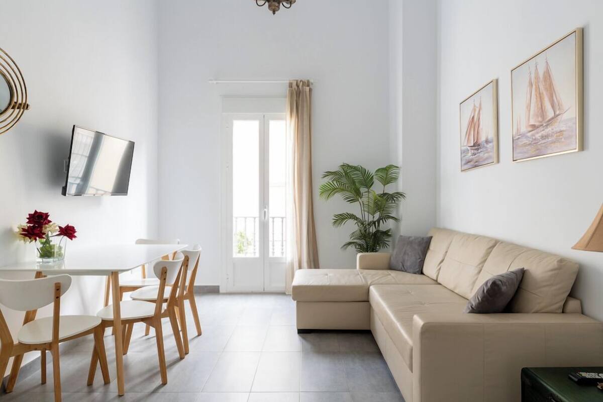 A modern living room with a beige sofa, a dining table with chairs, a TV mounted on the wall, and two framed paintings.