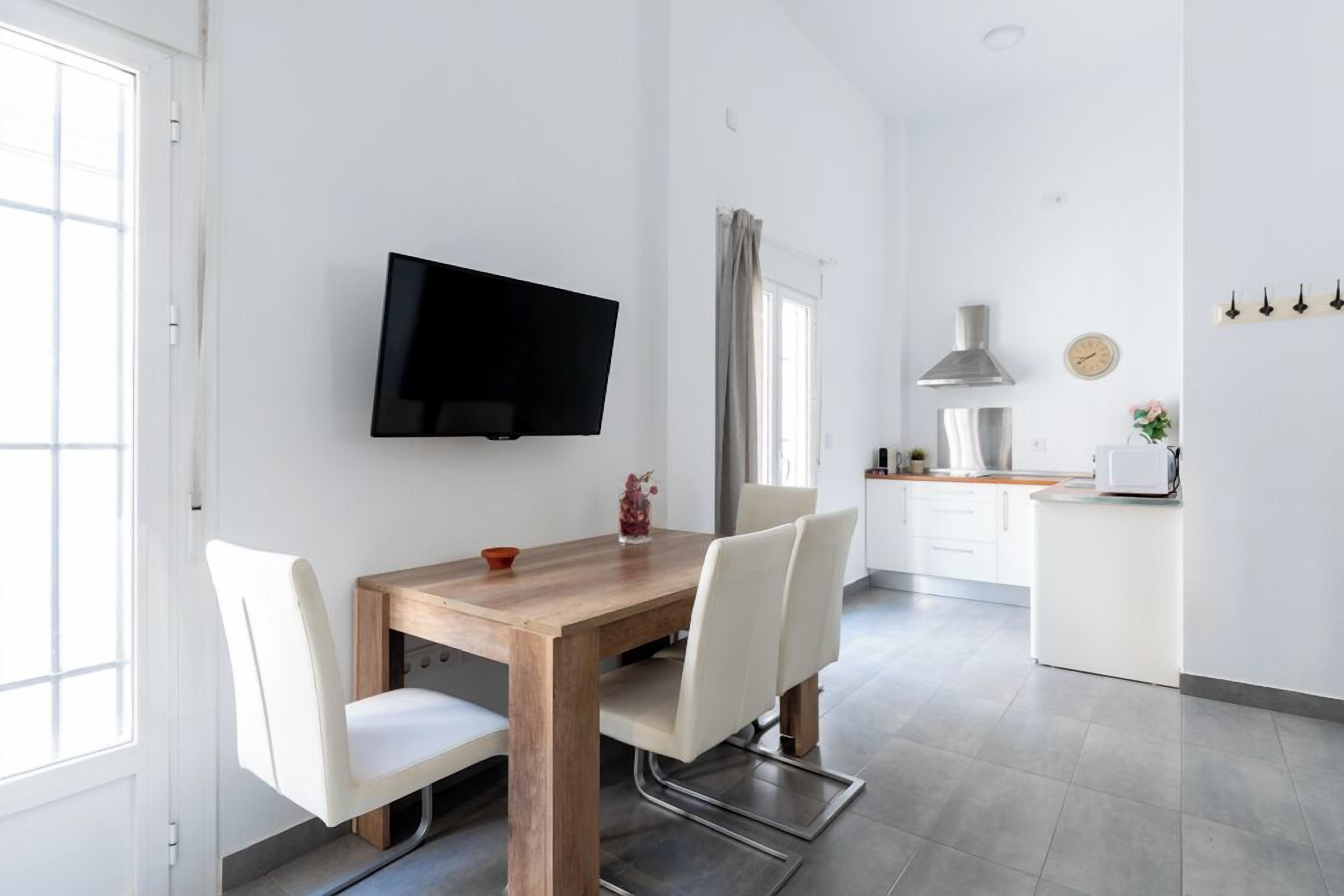 Une cuisine moderne avec une table à manger en bois, des chaises blanches, un téléviseur à écran plat fixé au mur et une fenêtre avec des rideaux.