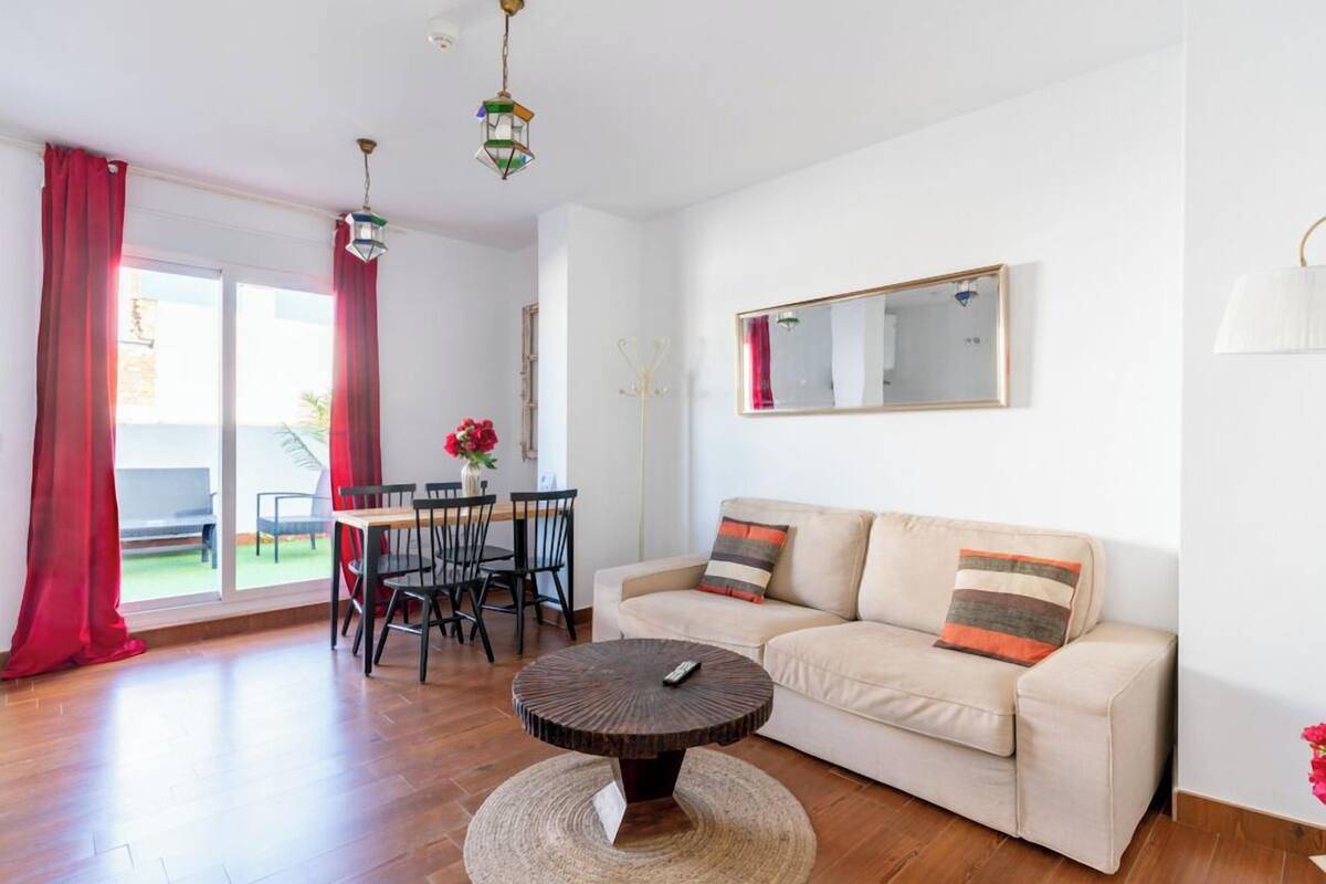 A living room with a sofa, round coffee table, dining table with chairs, and a large window with red curtains.