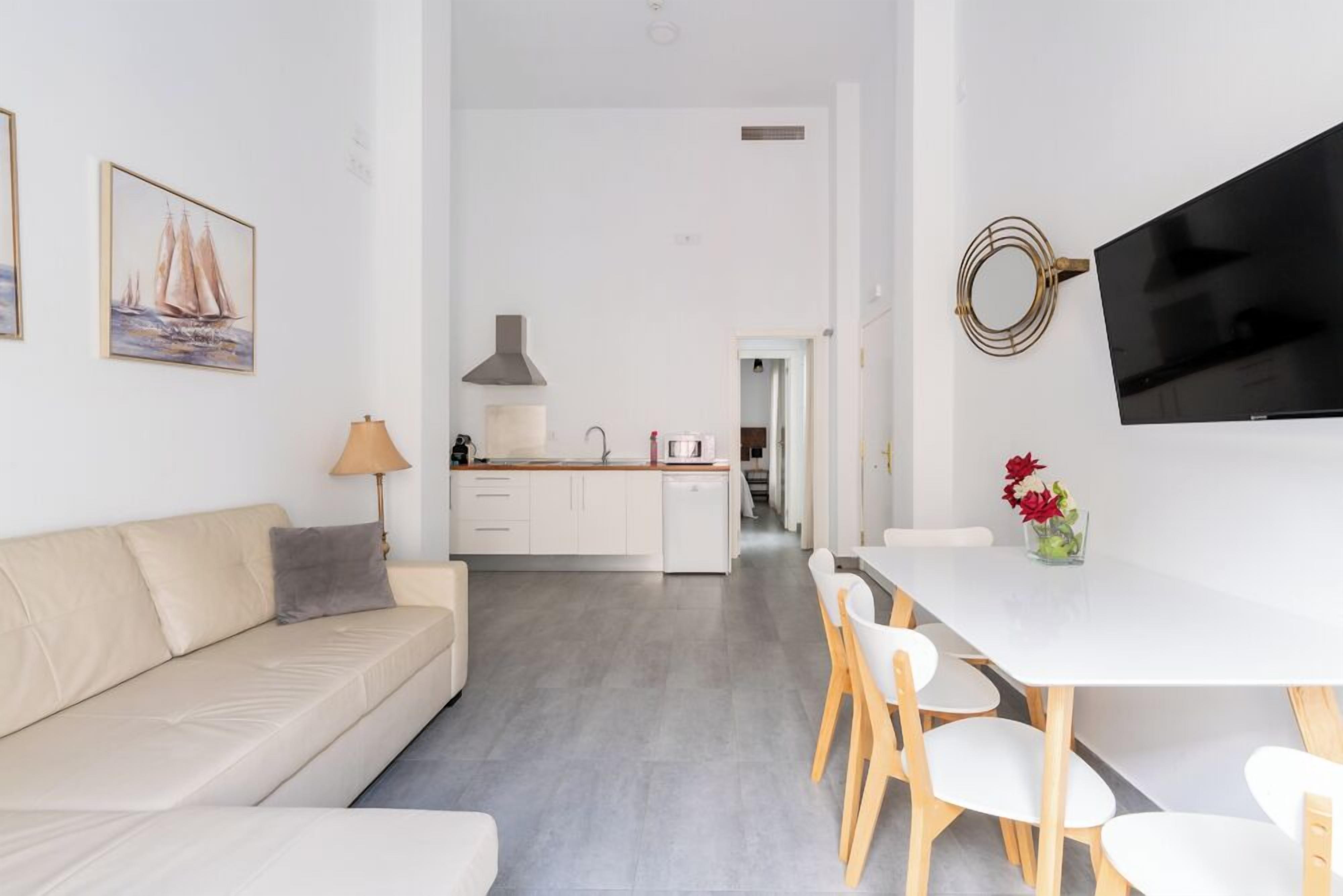 Una sala moderna con un sofá, una mesa de comedor con sillas, un televisor montado en la pared y una cocina al fondo.