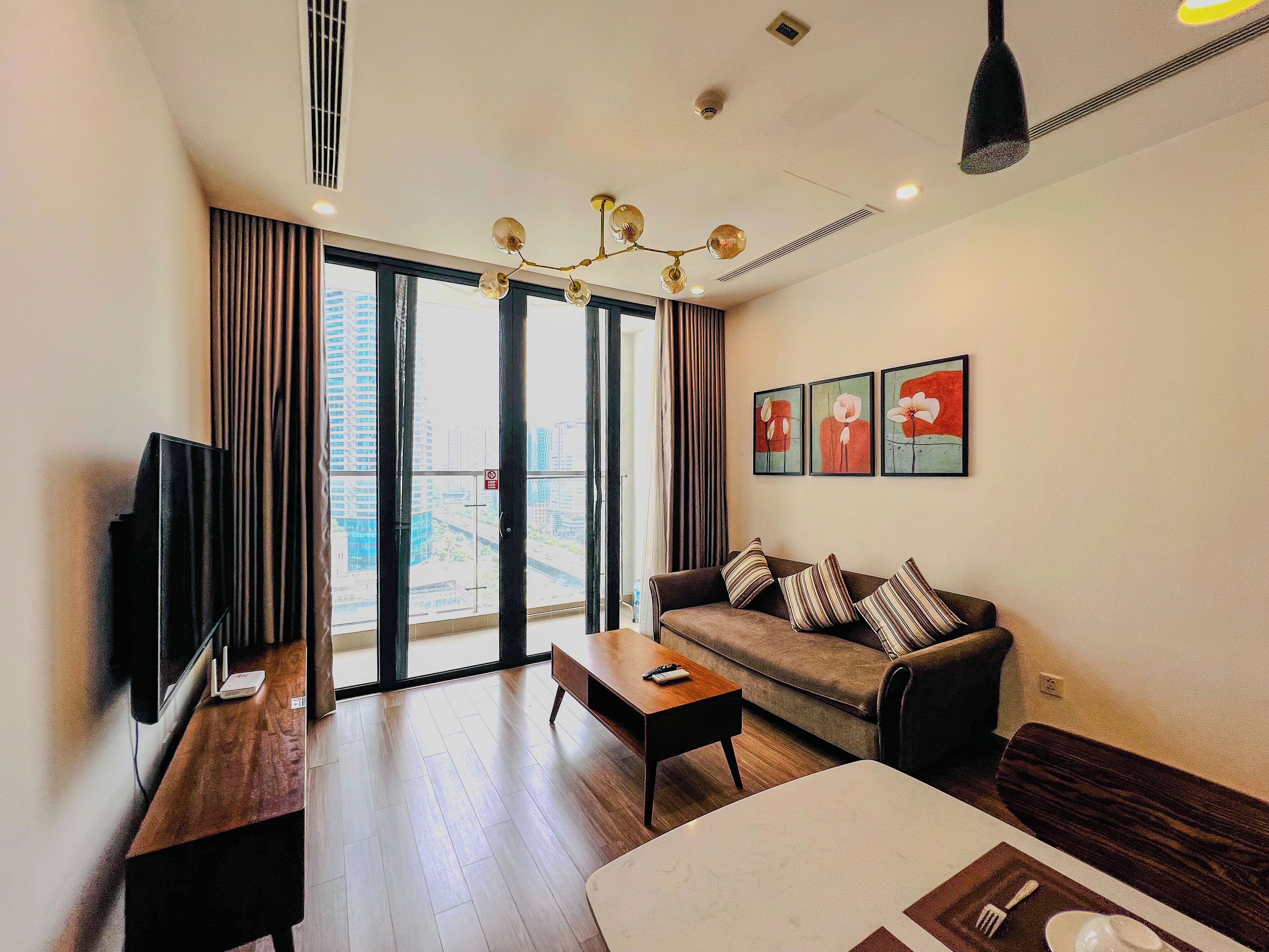 A modern living room with a sofa, coffee table, and flat-screen TV. Large windows offer a view of the cityscape.