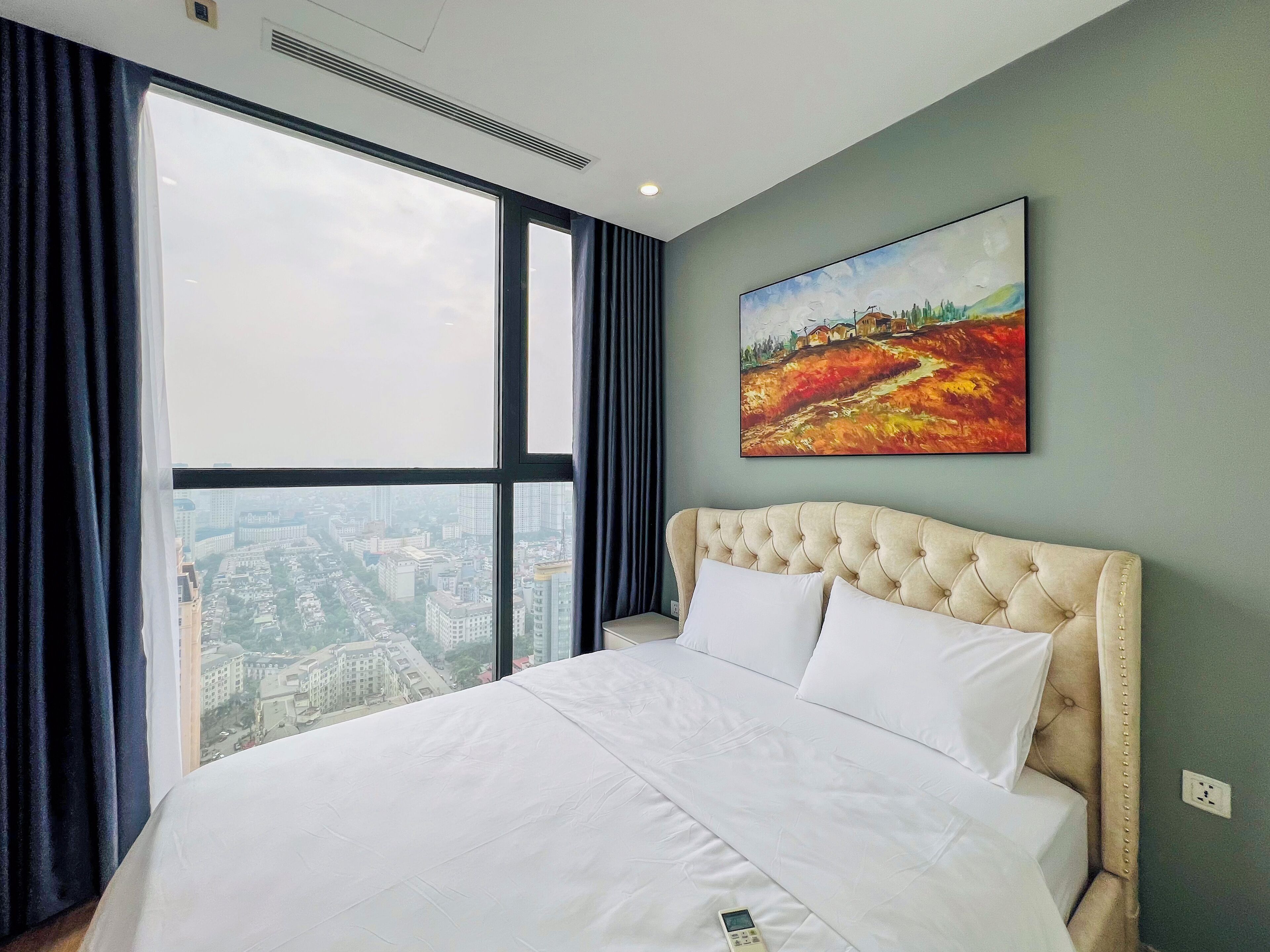 A hotel room with a large bed, a city view through the window, and a painting on the wall.