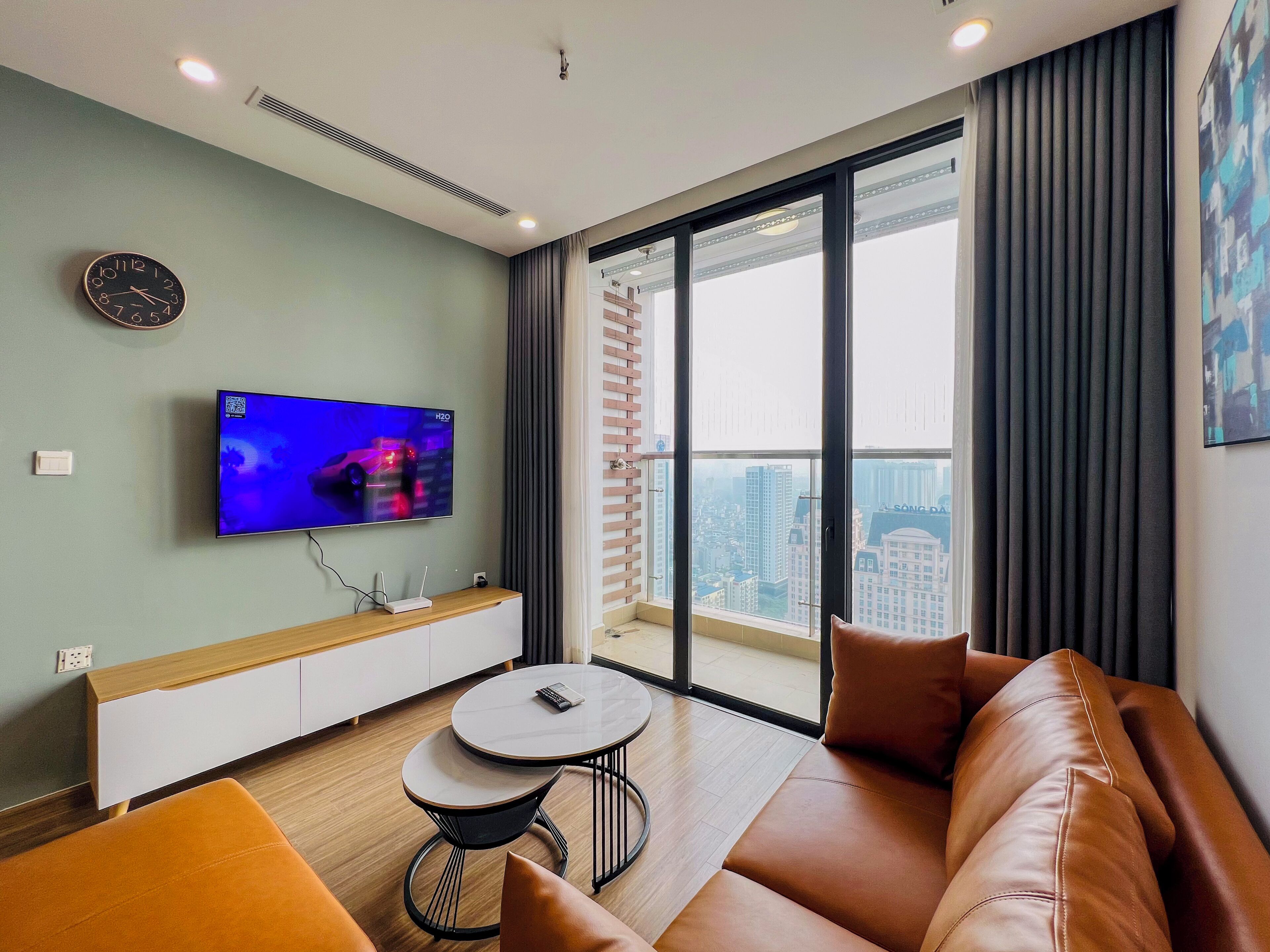 Ein modernes Wohnzimmer mit Flachbildfernseher, einer Couch, einem kleinen runden Tisch und Blick auf eine Stadtlandschaft durch große Fenster.