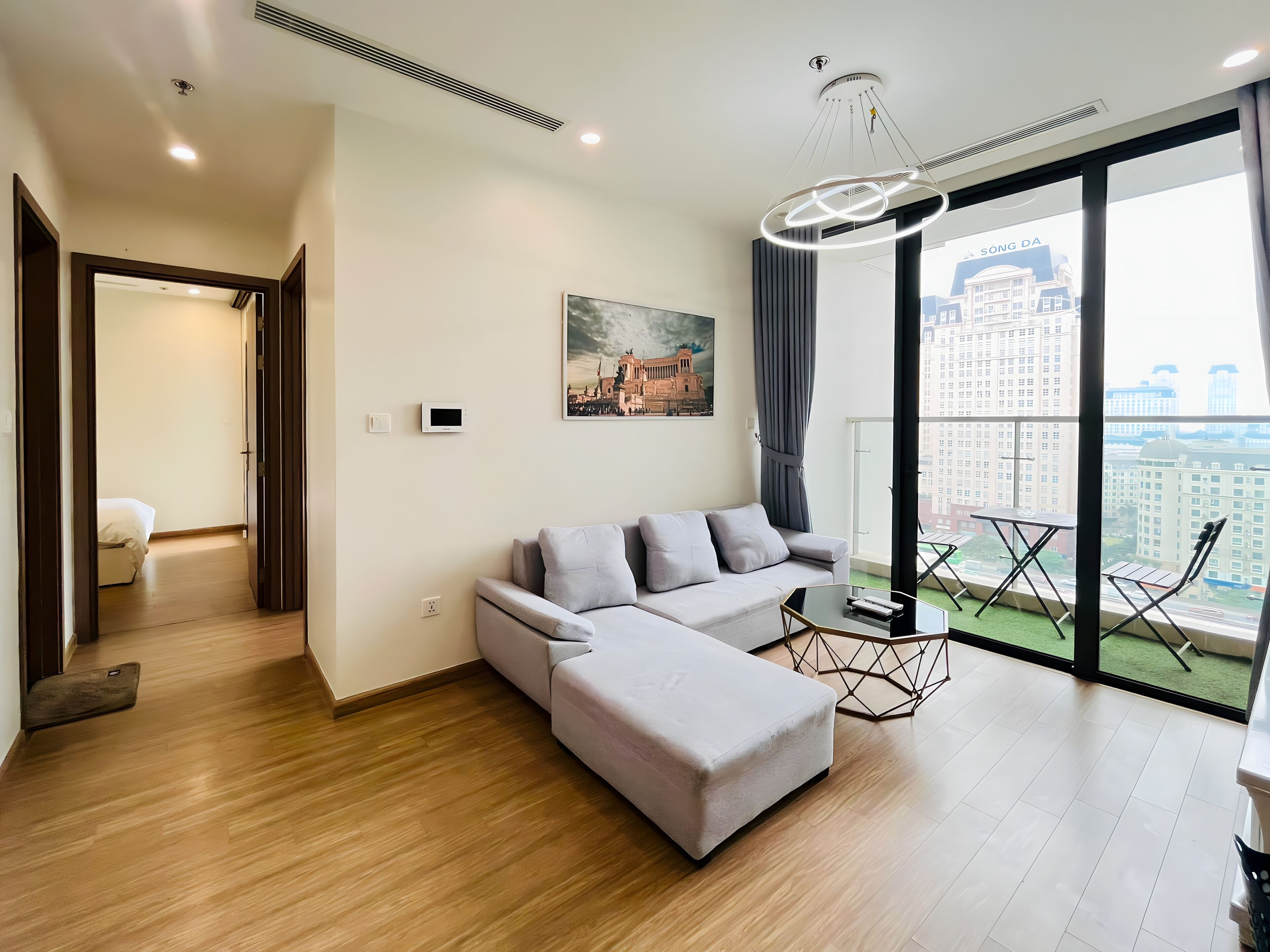 A modern living room with a grey sofa, a small table, and a view of a cityscape through large windows.