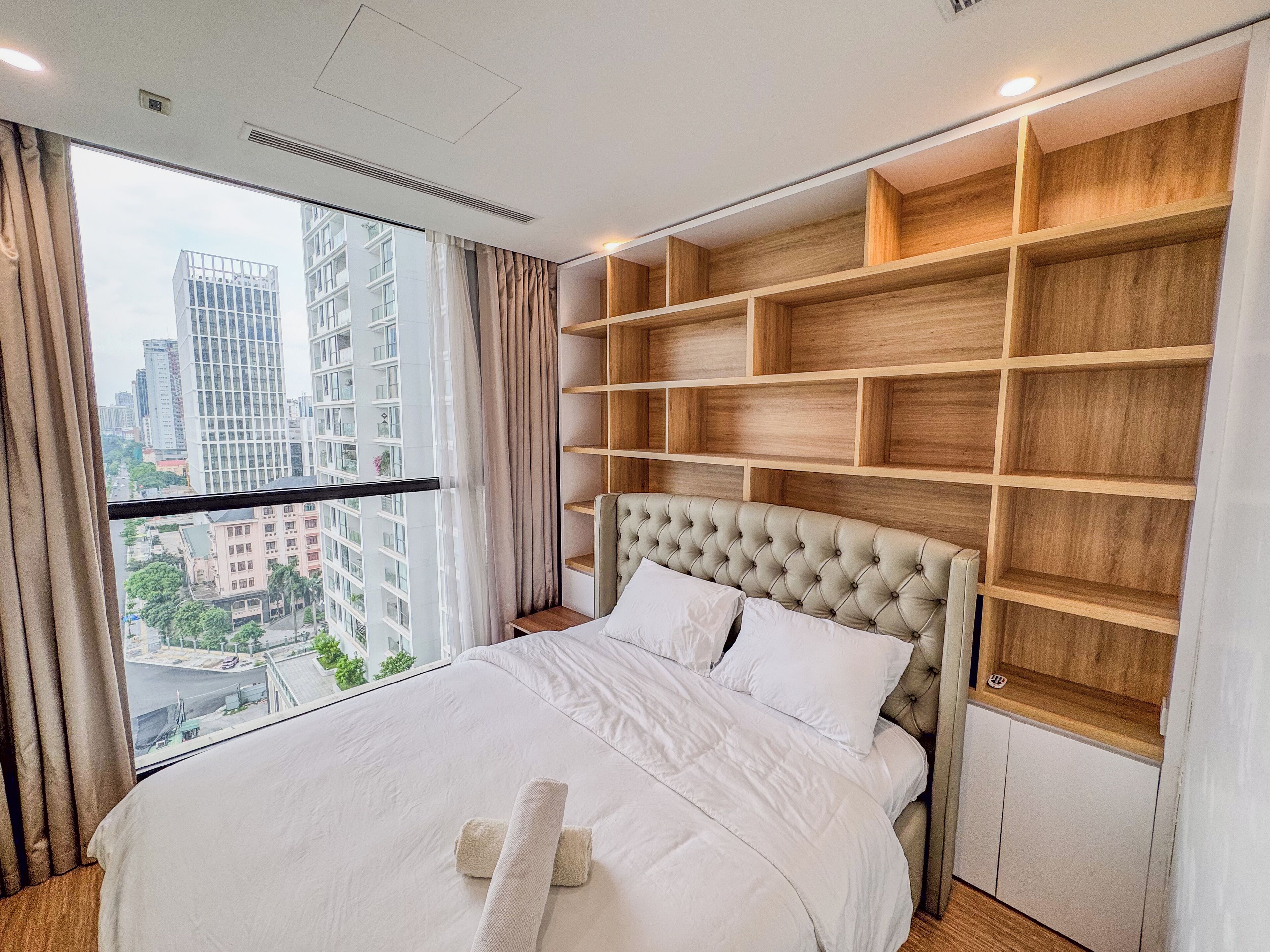A hotel room with a large bed, a view of the cityscape through the window, and built-in wooden shelves.