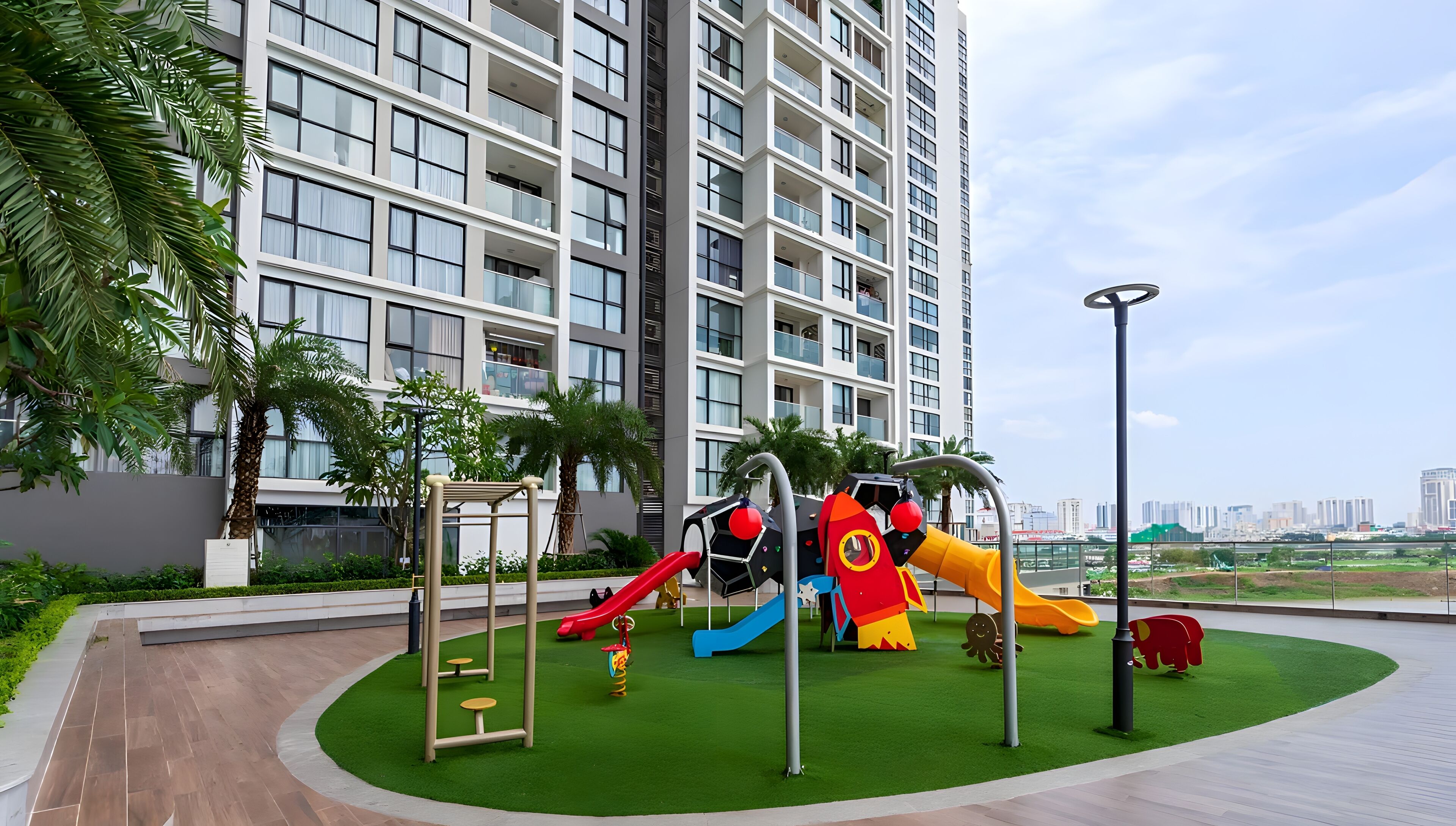 A playground with slides and climbing structures, surrounded by a landscaped area with palm trees, in front of a tall residential building.