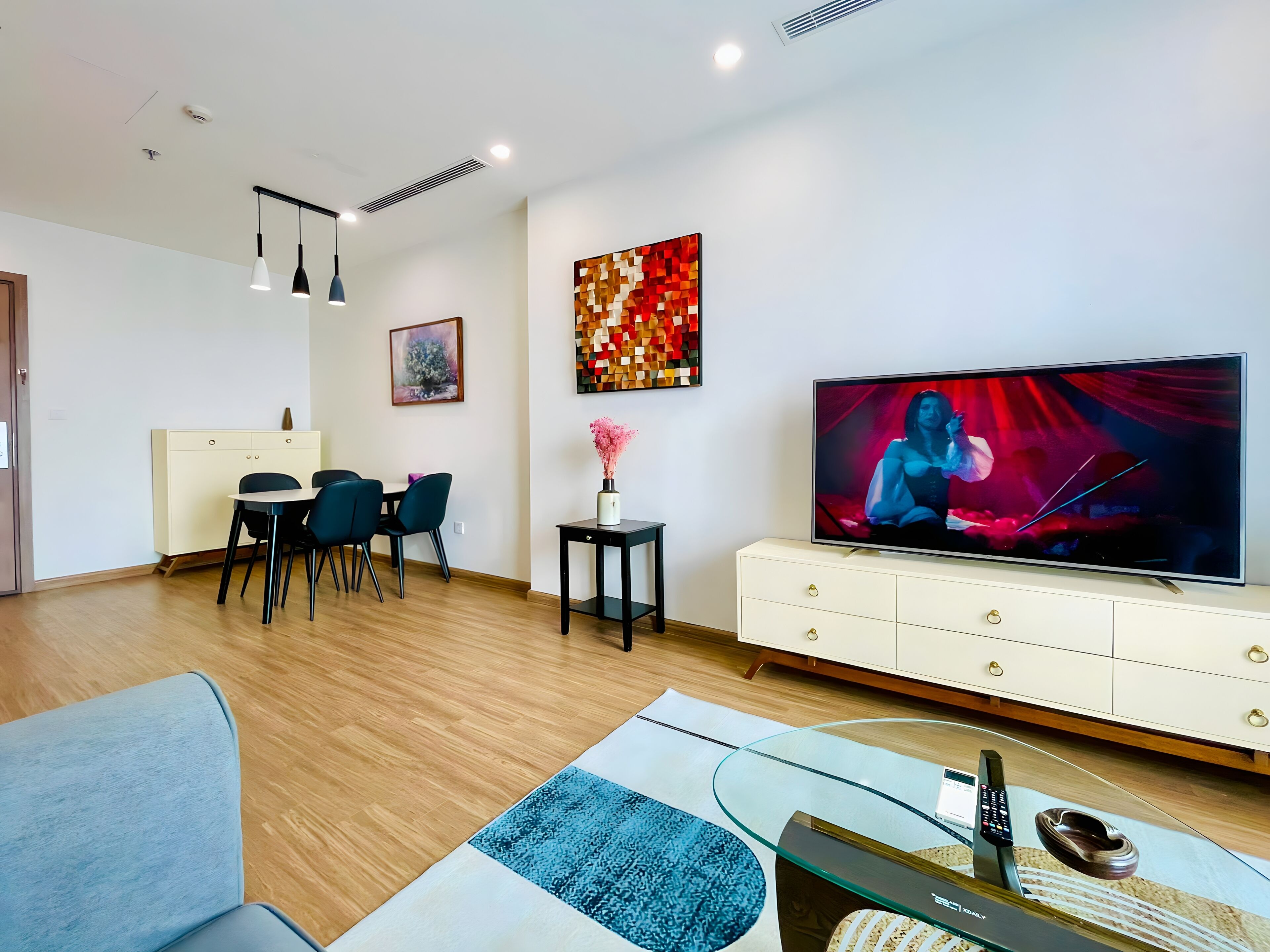 A modern living room with a flat-screen TV, a dining area with chairs, a coffee table, and a decorative vase.