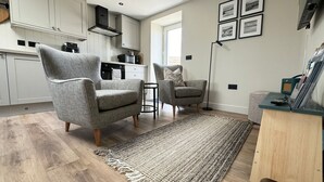 Una sala moderna con dos sillones grises, una mesita auxiliar, un tapete tejido y una cocina al fondo.