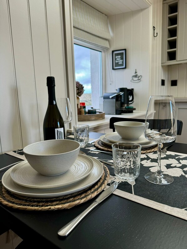 A dining table set with white plates, bowls, and clear glassware, with a bottle of wine and a coffee machine in the background. - No. 21 Beautiful & Cosy Modern Traditional Cottage (Isle of Lewis)
