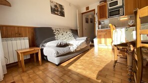 A cozy room with a bed, a small wooden table, a kitchen area with a microwave and oven, and a wooden chair.