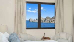 Ein Hotelzimmer mit einem Bett, zwei Stühlen, einem kleinen Tisch mit einer Dekoration und einem großen Fenster mit Blick auf zerklüftete Klippen und das Meer.