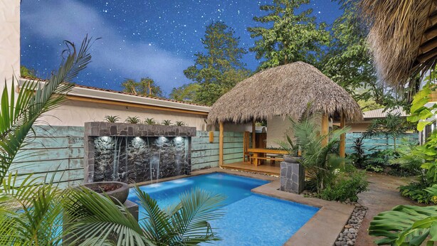 A swimming pool surrounded by tropical plants, a stone wall with a waterfall, and a thatched-roof structure. - Modern Home with Chic Bohemian Style and “Zen” Outdoor Oasis with Swimming Pool and Tiki-Style Dining Space (Potrero)