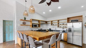 A modern kitchen with a central island, bar stools, a refrigerator, and pendant lights. - Modern Home with Chic Bohemian Style and “Zen” Outdoor Oasis with Swimming Pool and Tiki-Style Dining Space (Potrero)