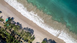 En vy över en strand sedd från luften, med kristallklart turkost vatten, en sandstrand och en rad med palmer.