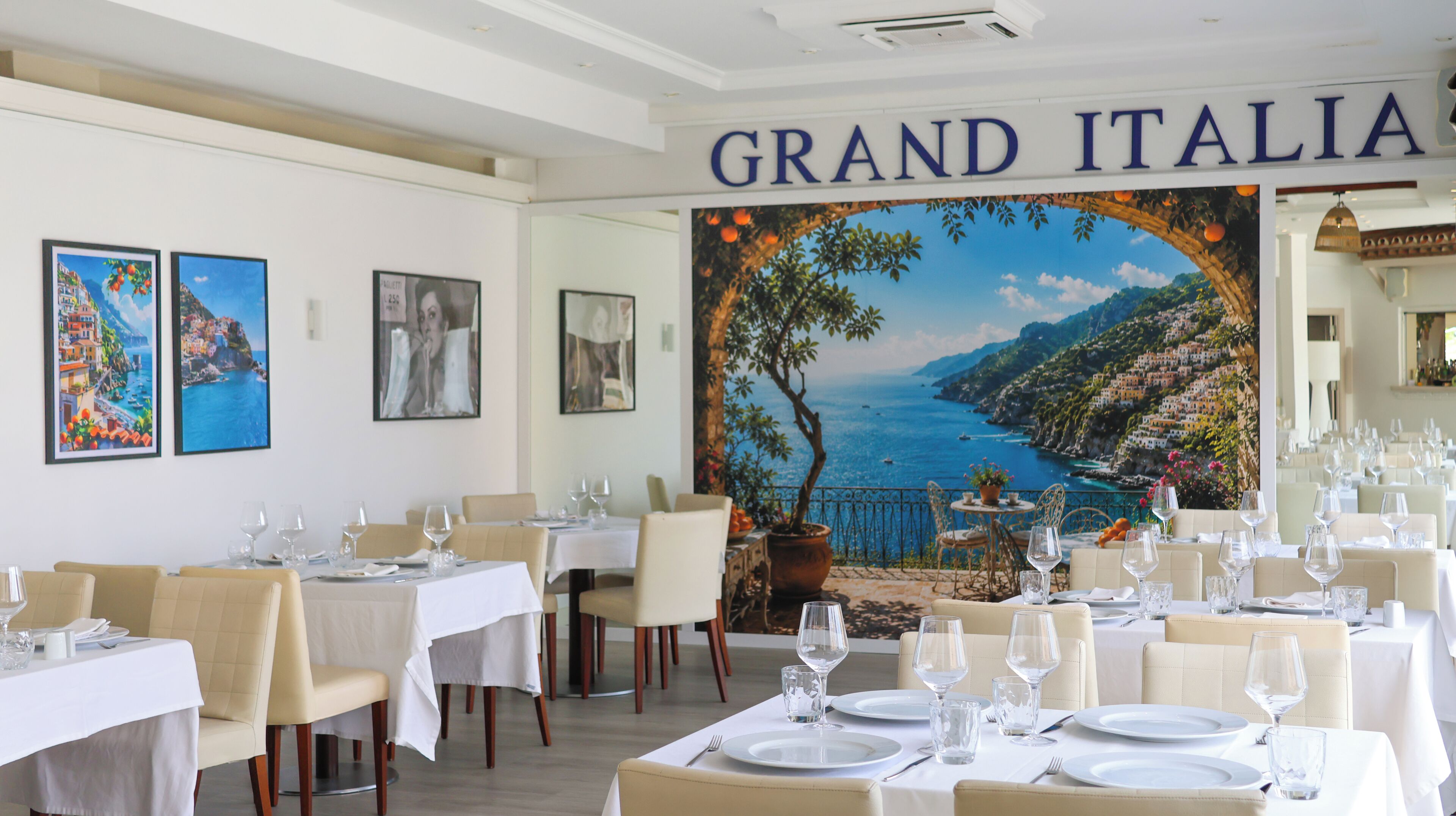 Un restaurant avec des tables dressées pour le service, une fresque représentant un paysage côtier, et l’enseigne « GRAND ITALIA » au-dessus de l’entrée.