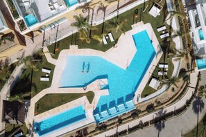 Vista aérea de un hotel moderno con una gran piscina, tumbonas y palmeras.