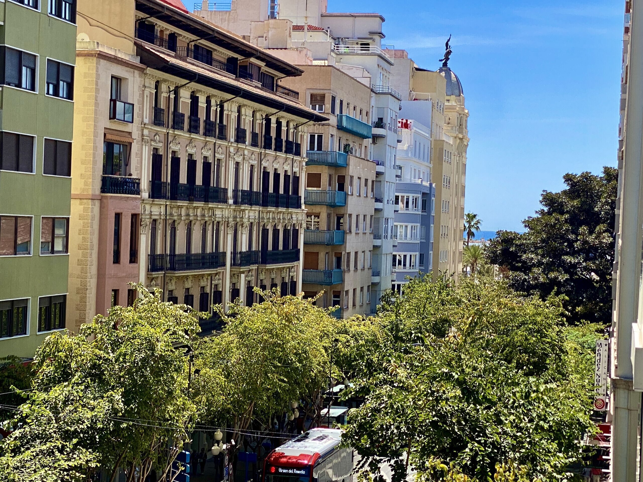 Un immeuble à plusieurs étages doté de balcons, un autobus rouge et de luxuriants arbres verts au premier plan.