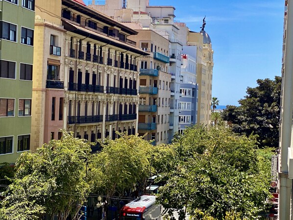 A multi-story building with balconies, a red bus, and lush green trees in the foreground.