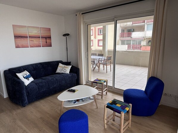 A living room with a dark blue sofa, a white coffee table, a blue armchair, and a colorful stool. A large window with a view of buildings is visible.