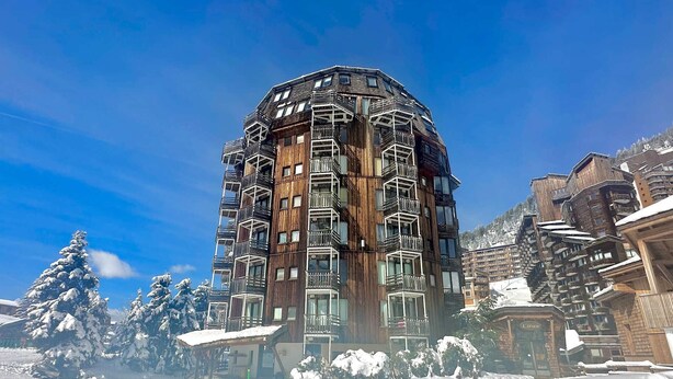Ein mehrstöckiges Holzgebäude mit Balkonen, umgeben von schneebedeckten Bäumen und blauem Himmel.