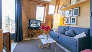 Ein gemütliches Wohnzimmer mit Fernseher, einer Couch, einem Couchtisch und einem Fenster mit Blick auf eine Holzterrasse.