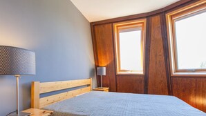 Ein Schlafzimmer mit einem hölzernen Bett, einem Nachttisch mit Lampe und zwei Fenstern.