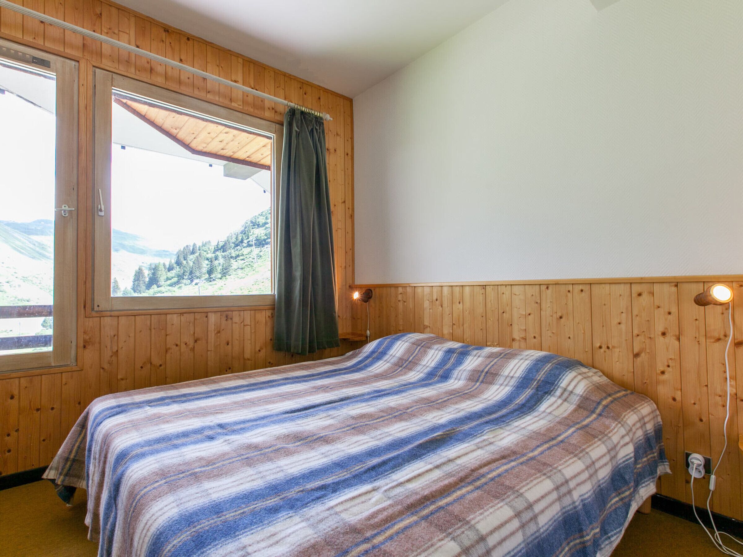 A room with wooden walls, a bed with a striped blanket, a window with a view of a forest, and a wall-mounted lamp.