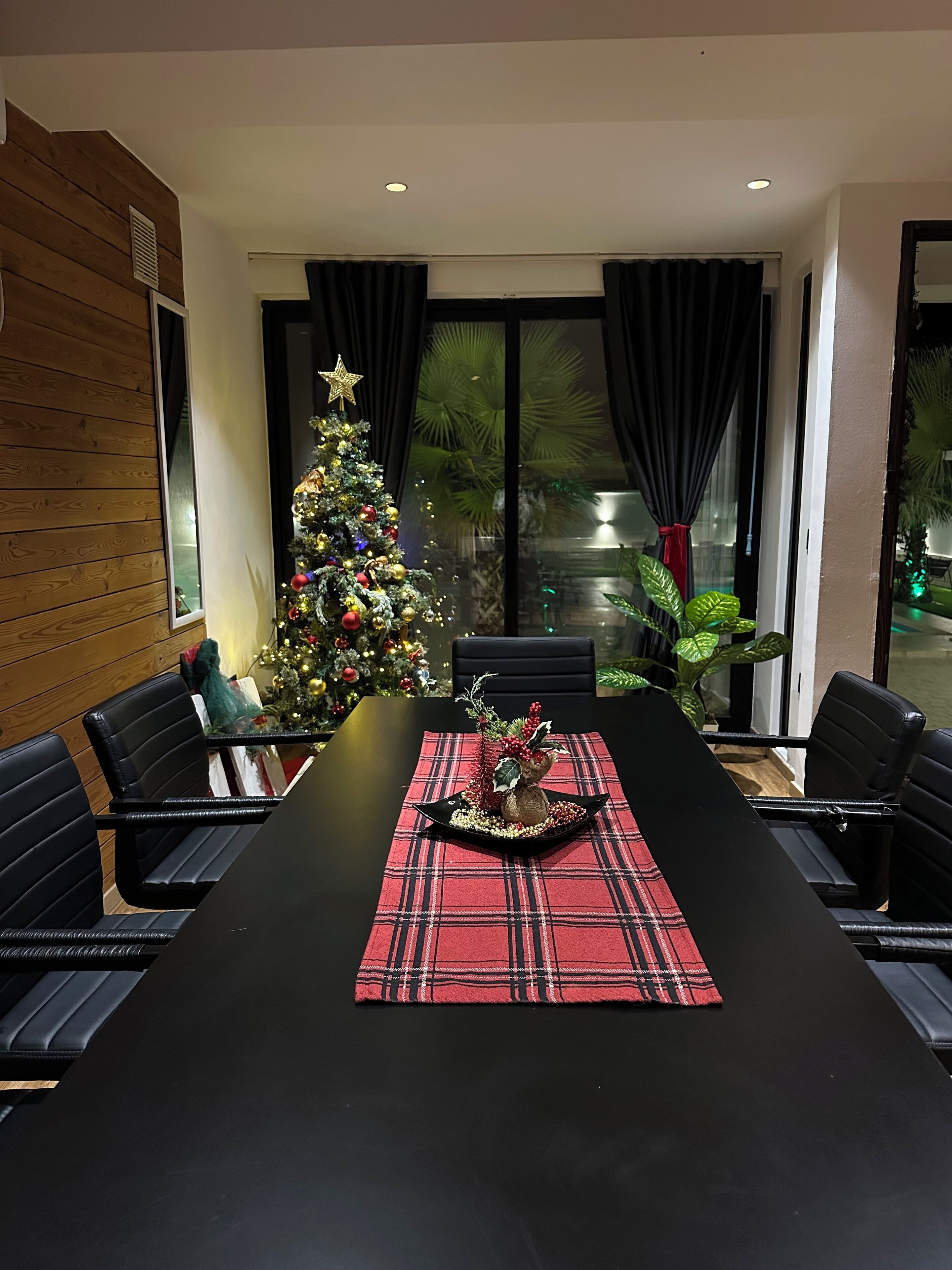 A modern dining area with a Christmas tree, black leather chairs, and a red plaid table runner.