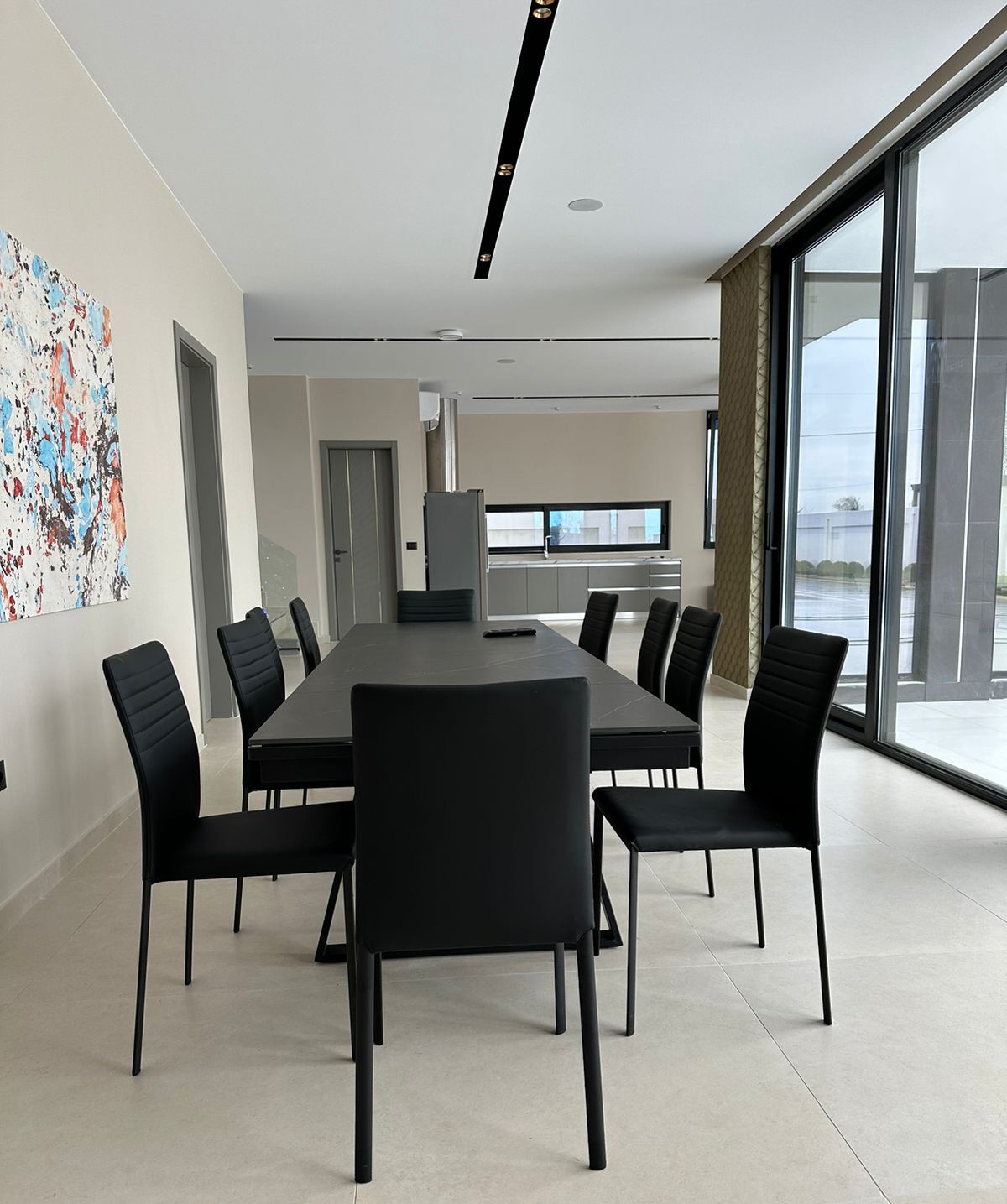 A modern meeting room with a large rectangular table and six black chairs, a large window with a view, and a kitchen area in the background.