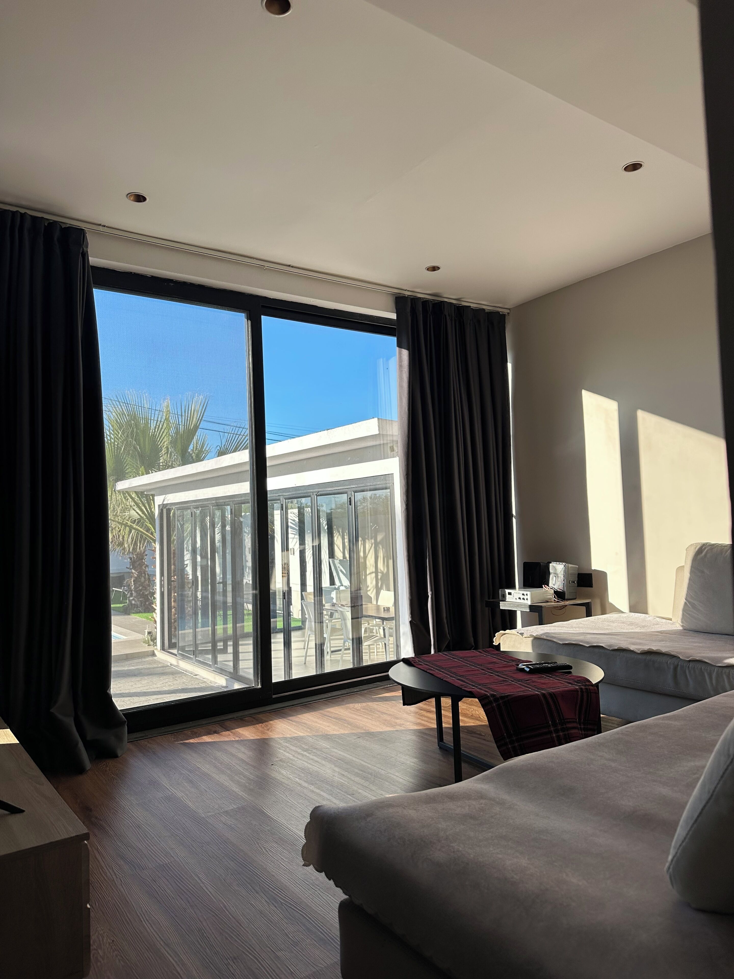 A modern hotel room with a large sliding glass door leading to a balcony, a bed, a small table with a remote control, and a TV on a side table.