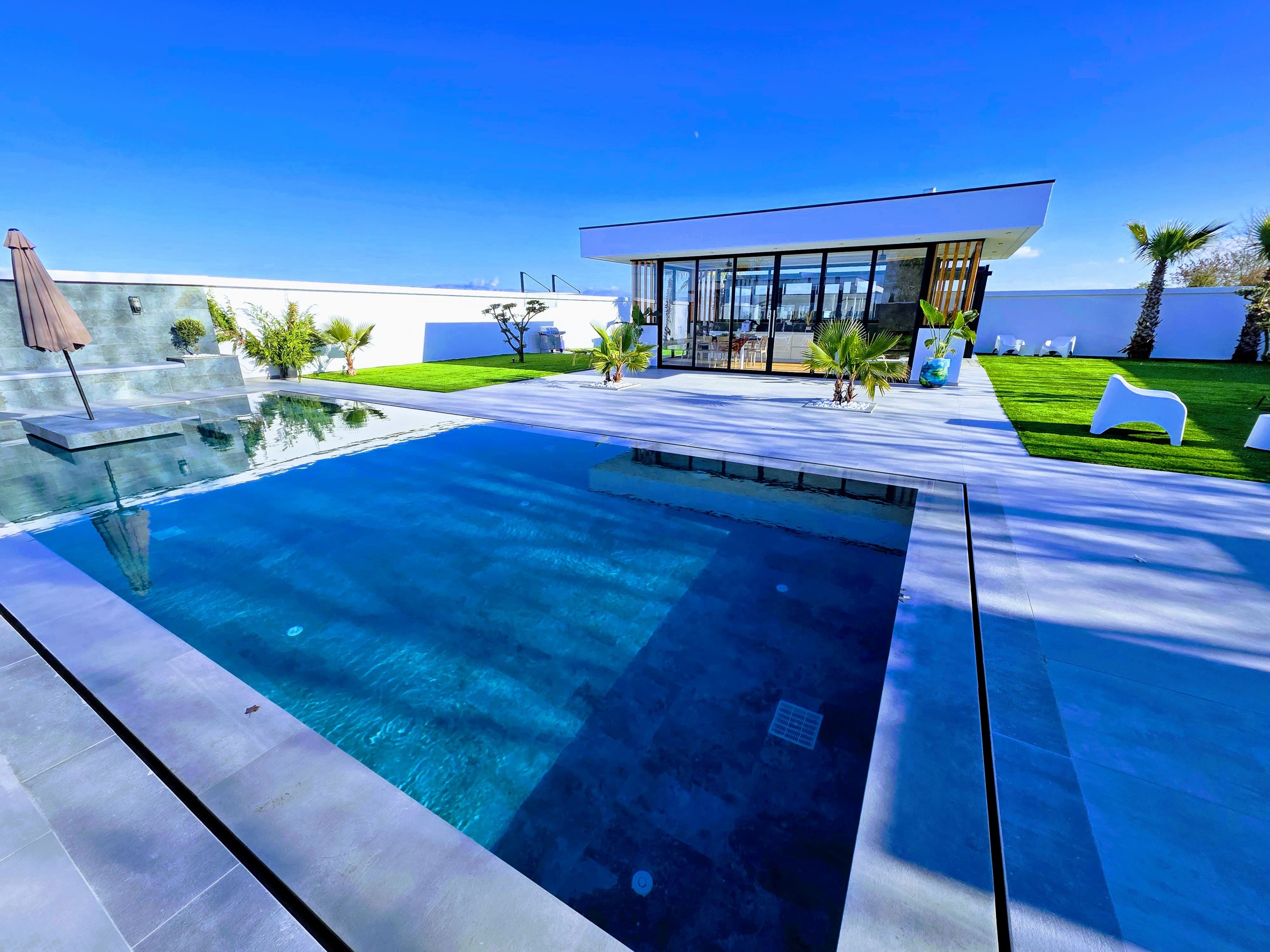 Un área de piscina moderna al aire libre con una piscina azul cristalina, una estructura de cristal y césped bien cuidado.